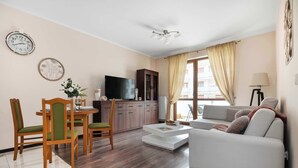 A living room with a sofa, a dining table with chairs, a TV, a cabinet, and a window with curtains. - Spacious Apartment with a Balcony | Parking | 2 Bedrooms (Toruń)