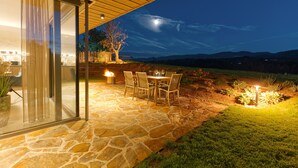 Patio esterno con pavimento in pietra, tavolo e sedie, un albero e cielo notturno con una luna visibile.