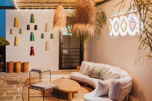 Um quarto aconchegante com um sofá branco curvo, uma mesa de centro redonda de madeira e uma pequena mesa preta de metal. Há plantas em vasos e uma parede decorativa com várias formas.