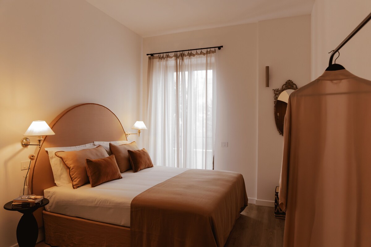 A bedroom with a bed, bedside lamps, a window with curtains, a coat on a hanger, and a small table with books.