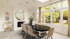 A dining room with a round table, six chairs, a gold-framed side table, a mirror, and a fireplace. - The Steele Blue House (London)