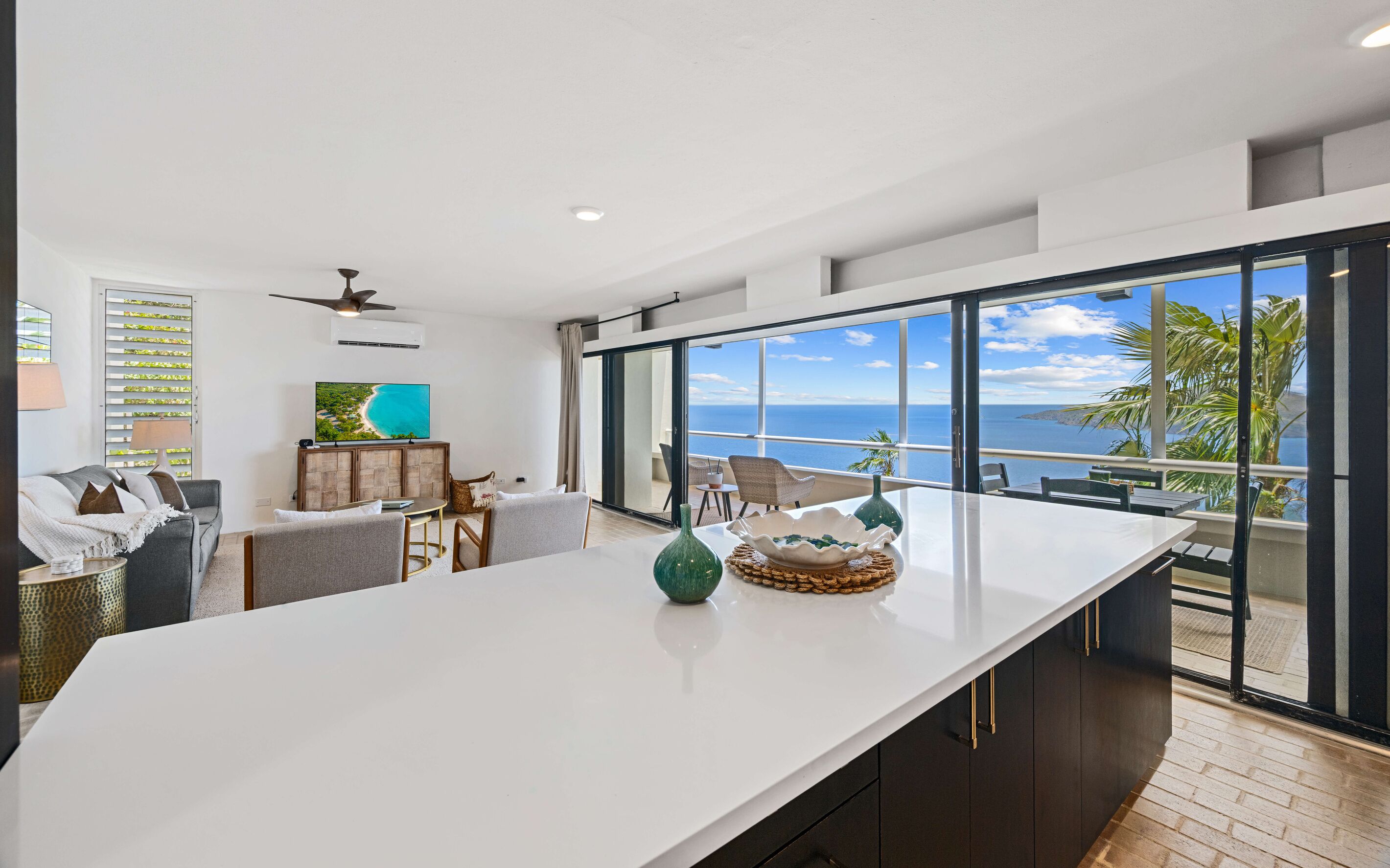Un salon moderne doté d’une grande fenêtre donnant sur la mer, d’un téléviseur à écran plat et d’un coin repas avec une table blanche.