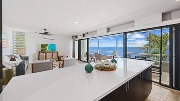 Un salon moderne doté d’une grande fenêtre donnant sur la mer, d’un téléviseur à écran plat et d’un coin repas avec une table blanche.