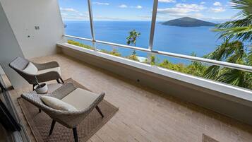 Un balcon avec des fauteuils en osier et une petite table, offrant une vue sur la côte, avec une montagne et la mer en toile de fond.