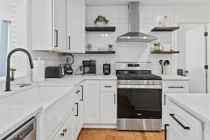 A modern kitchen with white cabinetry, a stainless steel stove, and a built-in oven. There are black countertops, a coffee maker, and a toaster. - The Tranquil Token-hot Tub & Ping Pong Table (North Charleston)