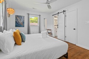 Ein Schlafzimmer mit einem großen Bett, weißer Bettwäsche, einem Deckenventilator, einem Fenster mit Vorhängen und einer Tür zu einem anderen Raum.