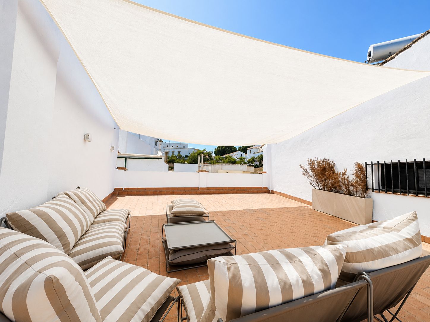 Un patio exterior con un toldo blanco, un área de estar con cojines a rayas y vistas de los edificios circundantes.