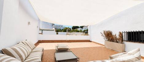 Un patio exterior con un toldo blanco, un área de estar con cojines a rayas y vistas de los edificios circundantes.