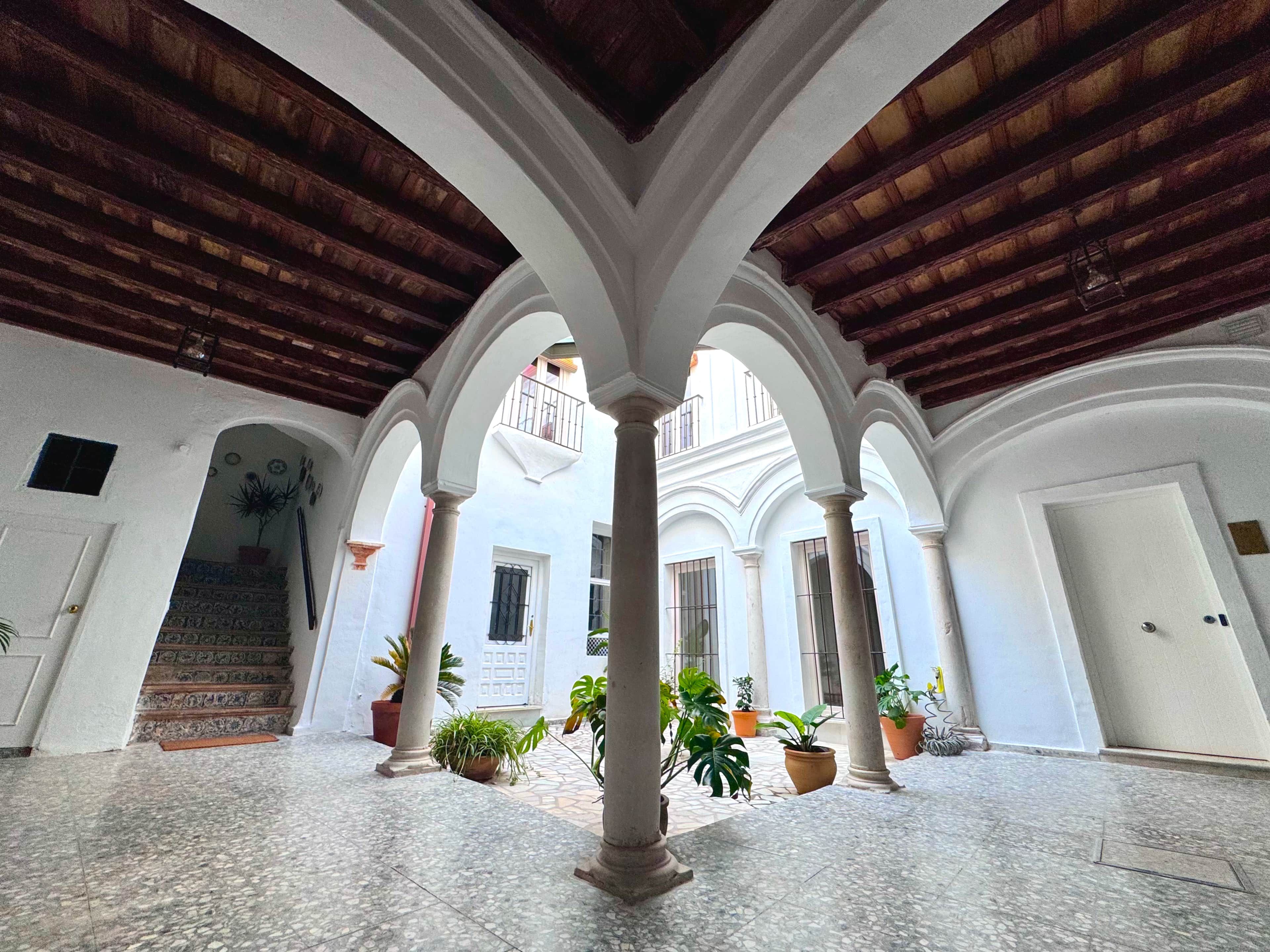 An interior courtyard with a tiled floor, white walls, and a wooden ceiling. There are columns supporting the structure, and potted plants are placed around the area.