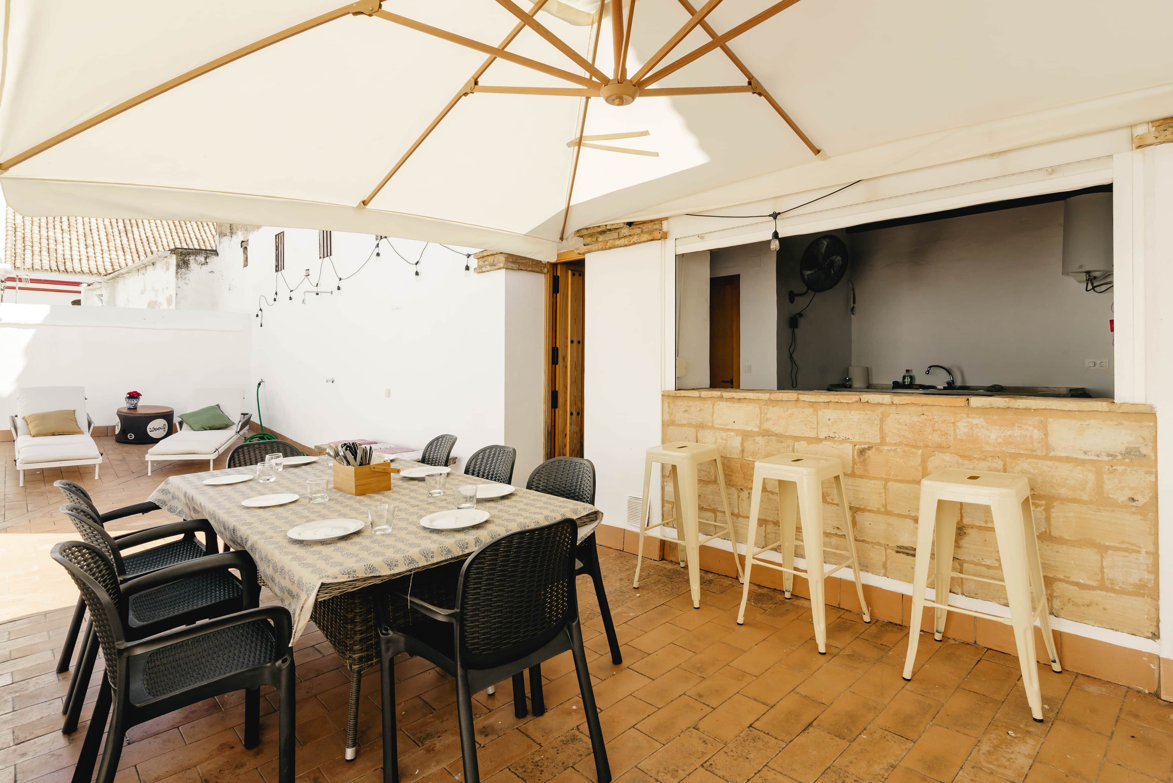 Un'area pranzo con un tavolo apparecchiato per quattro persone, circondato da sedie, una zona bar con sgabelli e una cucina visibile attraverso un'apertura.