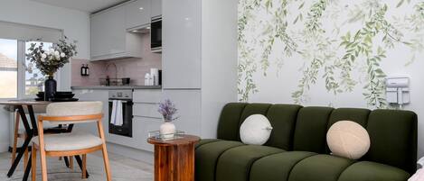 A modern kitchen with white cabinets, a dining area with a wooden chair, and a green sofa with decorative pillows.