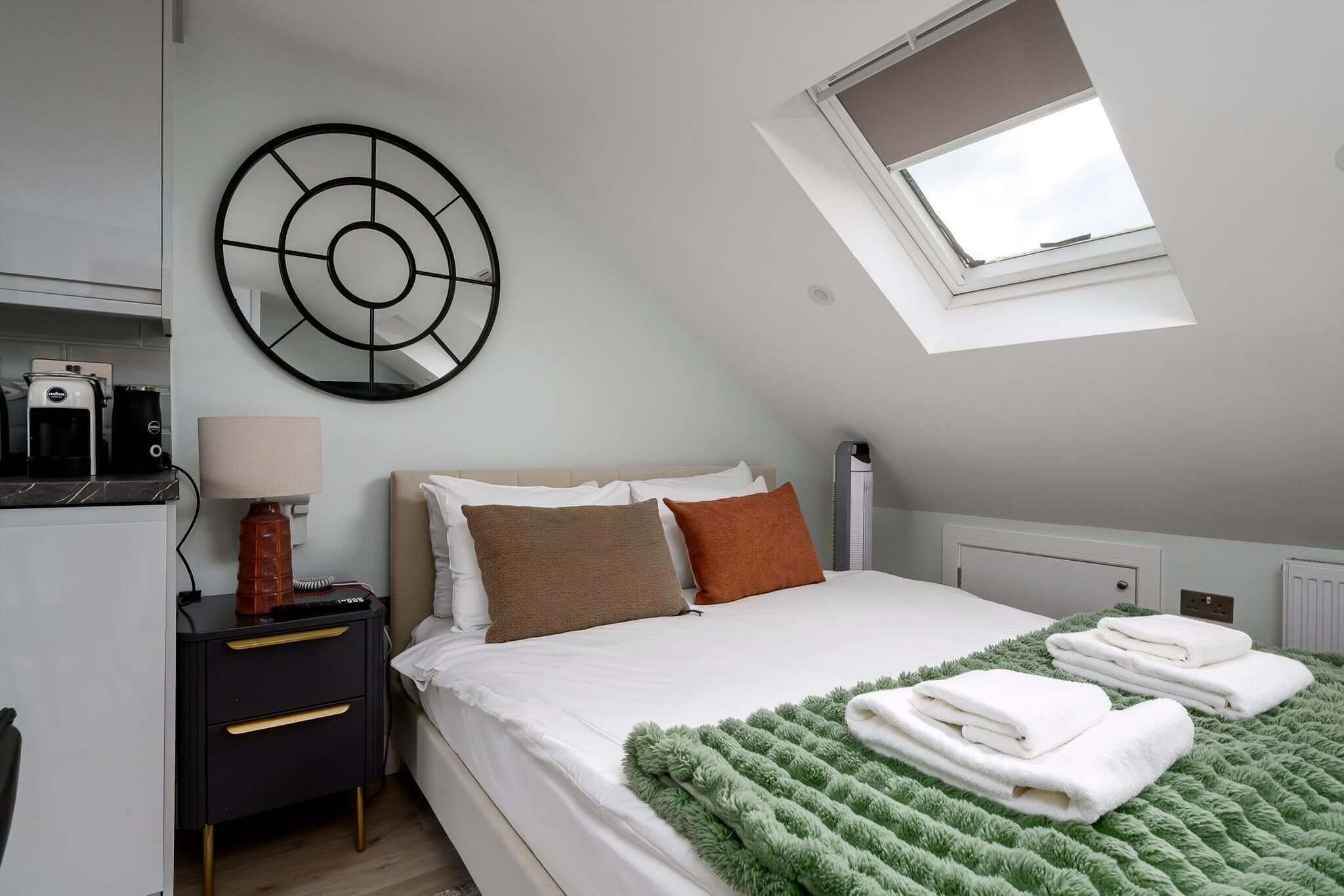 A bedroom with a skylight, a bed with white linens, a green blanket, and a wall clock.