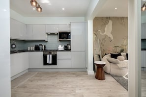 A modern kitchen with white cabinetry, a built-in oven, and a microwave. There is a wooden floor and a small round wooden table. - Greenford Apartments (Wembley)
