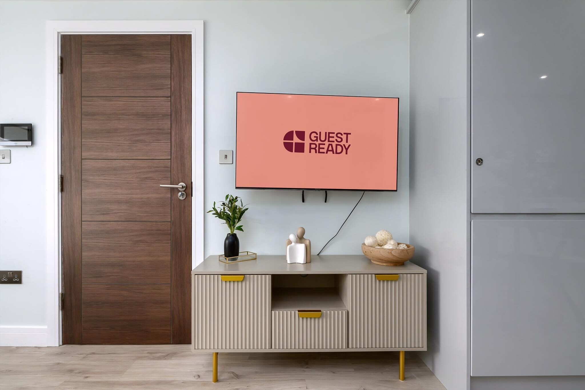 A modern hotel lobby with a wooden door, a grey console table with yellow handles, a mounted television displaying 'GUEST READY', and a small plant on the table.