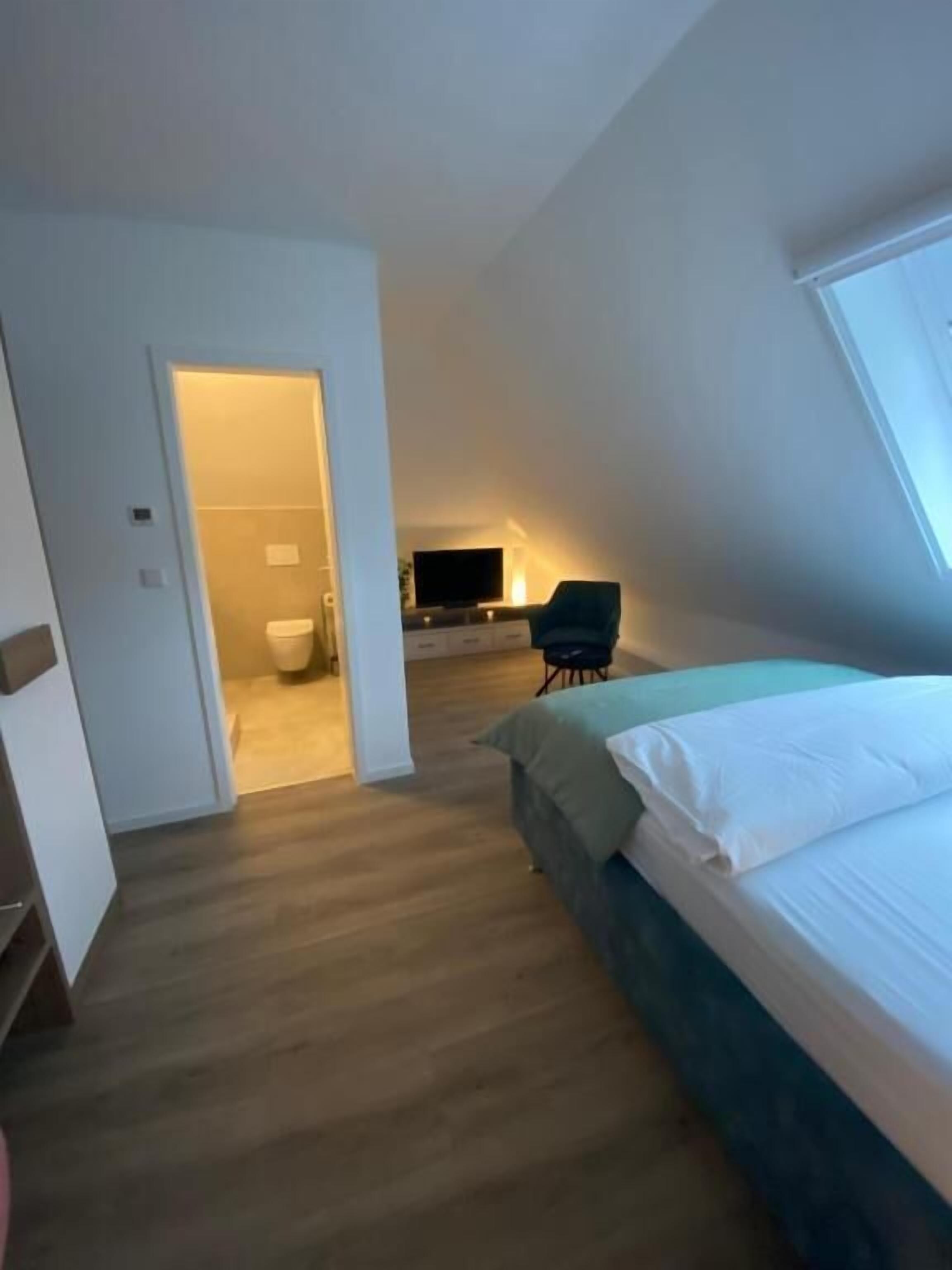 A bedroom with a bed, a chair, and a television in the background.