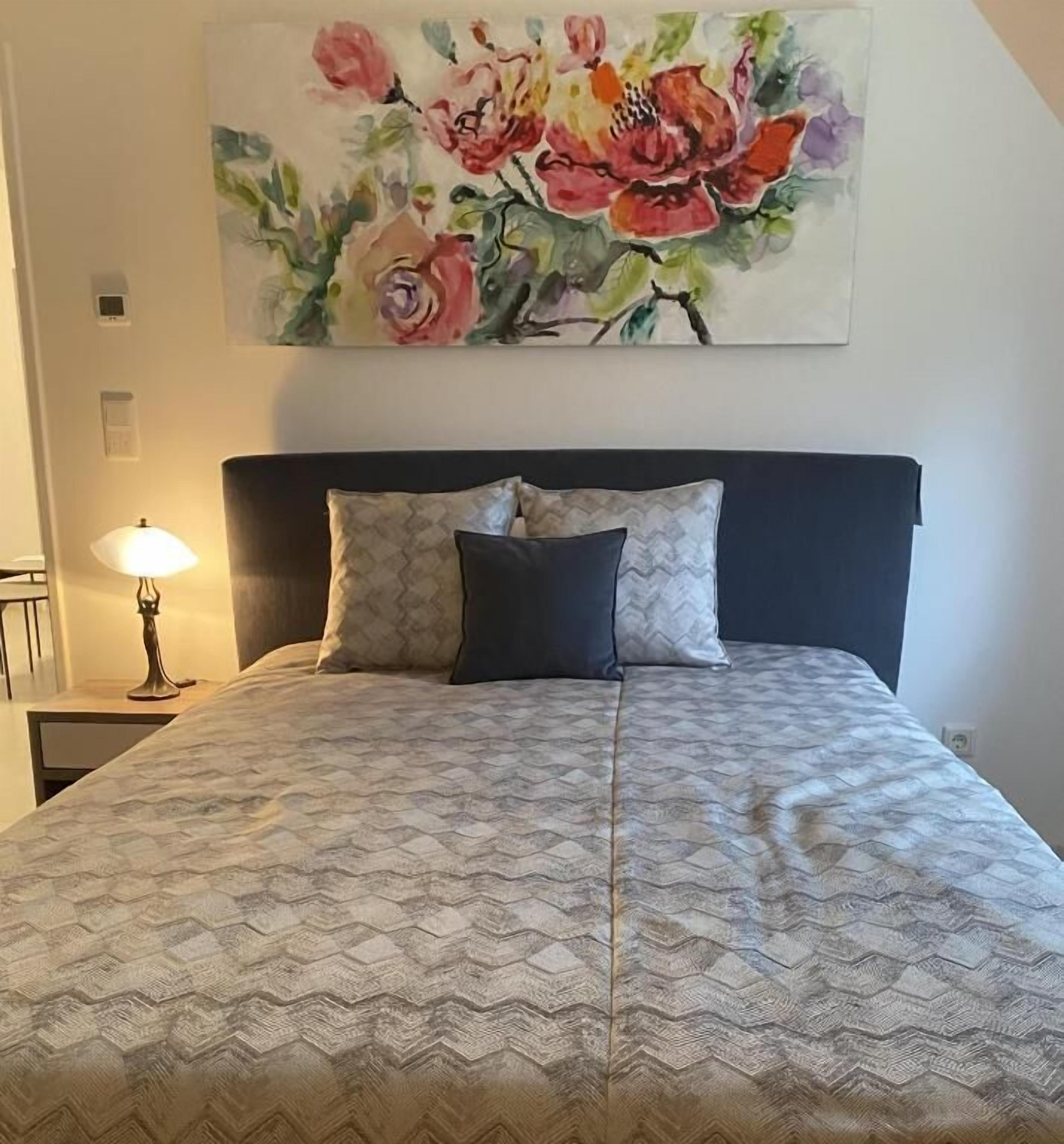 Uma cama com colcha estampada, duas almofadas e um quadro de flores pendurado na parede.