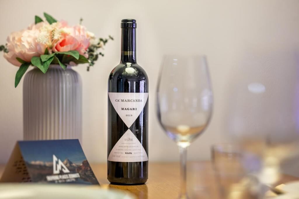 A wine bottle labeled 'CA'MARCANDA MAGARI 2019' on a table with a glass and a vase of flowers.
