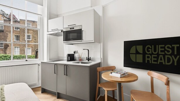 A modern kitchen with a built-in oven and microwave, a small dining area with two chairs, and a window with a view of buildings. - Guestready - The Crescent (London)