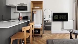 A modern kitchen with a built-in oven, microwave, and coffee maker. There is a wooden chair and a small table, and a 'GUEST READY' sign on the wall. - Guestready - The Crescent (London)