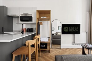 A modern kitchen with a built-in oven, microwave, and coffee maker. There is a wooden chair and a small table, and a 'GUEST READY' sign on the wall.