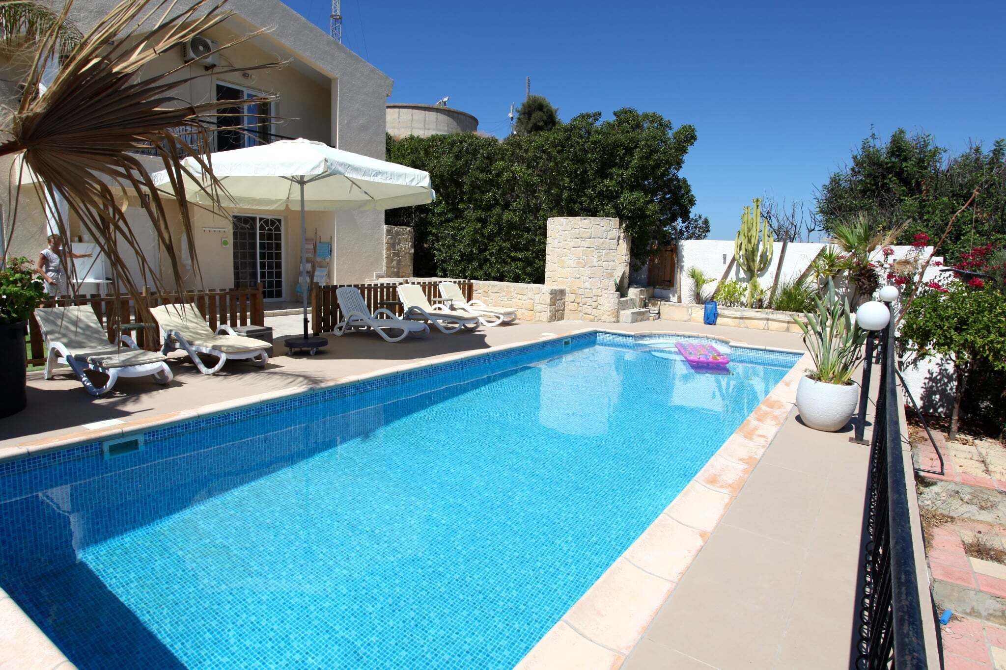 A swimming pool with lounge chairs and umbrellas, surrounded by greenery and a stone wall.