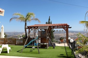 A playground with a slide and swings, surrounded by a wooden fence, palm trees, and a view of distant hills. - Stunning 5-bedroom villa with private pool, panoramic sea and mountain views. (Pissouri)