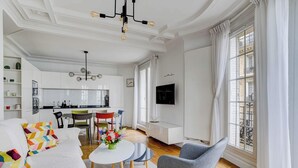 Ett modernt vardagsrum med en soffa, ett matbord och stolar, ett kök i bakgrunden och stora fönster med gardiner.