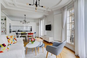 A modern living room with a sofa, dining table, and chairs, a kitchen in the background, and large windows with curtains. - Charming Eiffel Stylish 1 BR With Parisian Charm (Paris)