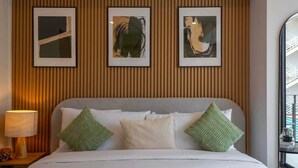 Hotellihuone, jossa on sänky, tyynyjä, lamppu ja kolme kehystettyä taideteosta seinällä.