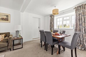 A dining area with a wooden table, grey upholstered chairs, a side table with a lamp, and patterned curtains. - Cosy Cottage in Rural Location With Lovely Country (Malton)