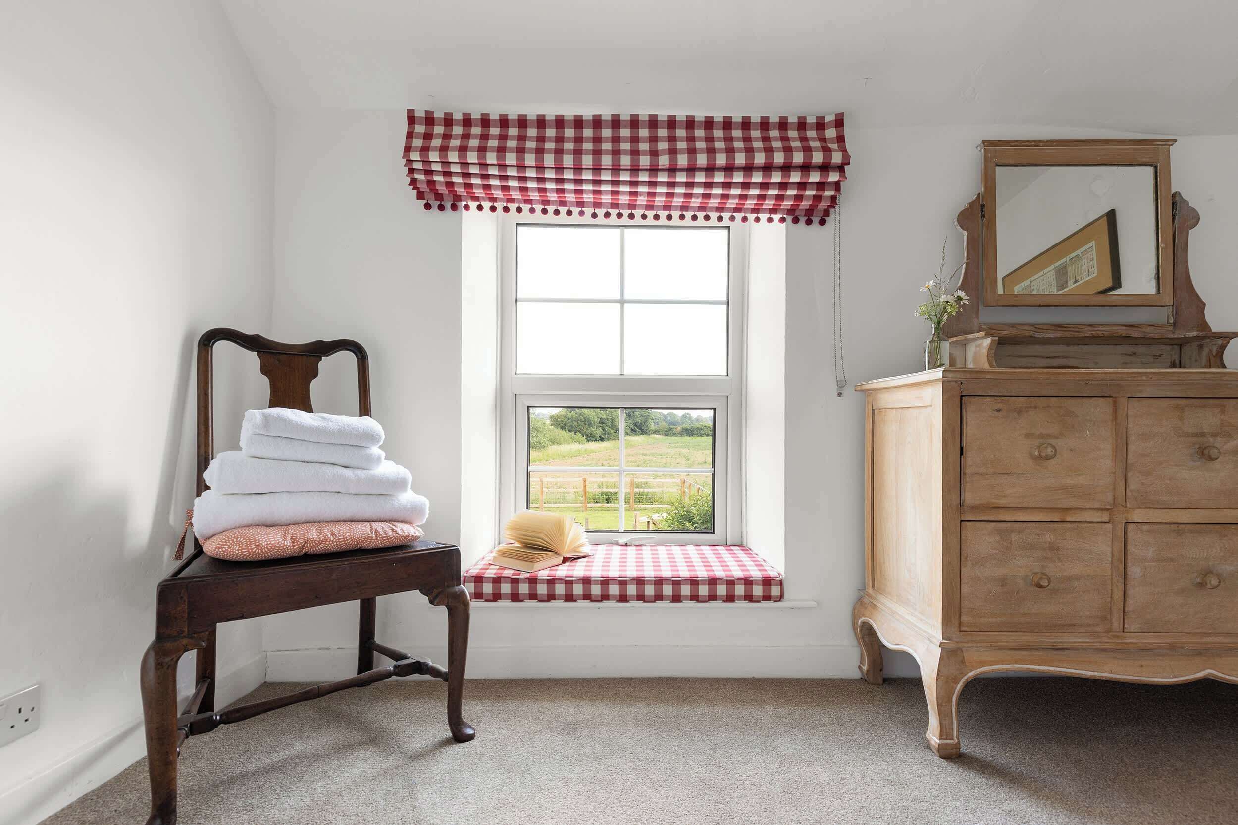 A room with a window, a wooden chair with towels, a wooden dresser, and a checkered valance.