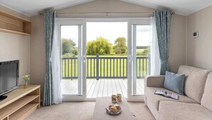 Ein Wohnzimmer mit Fernseher, Sofa und Couchtisch, ein Balkon mit Holzterrasse und Blick ins Grüne.