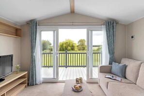 Ein Wohnzimmer mit Fernseher, Sofa und Couchtisch, ein Balkon mit Holzterrasse und Blick ins Grüne.