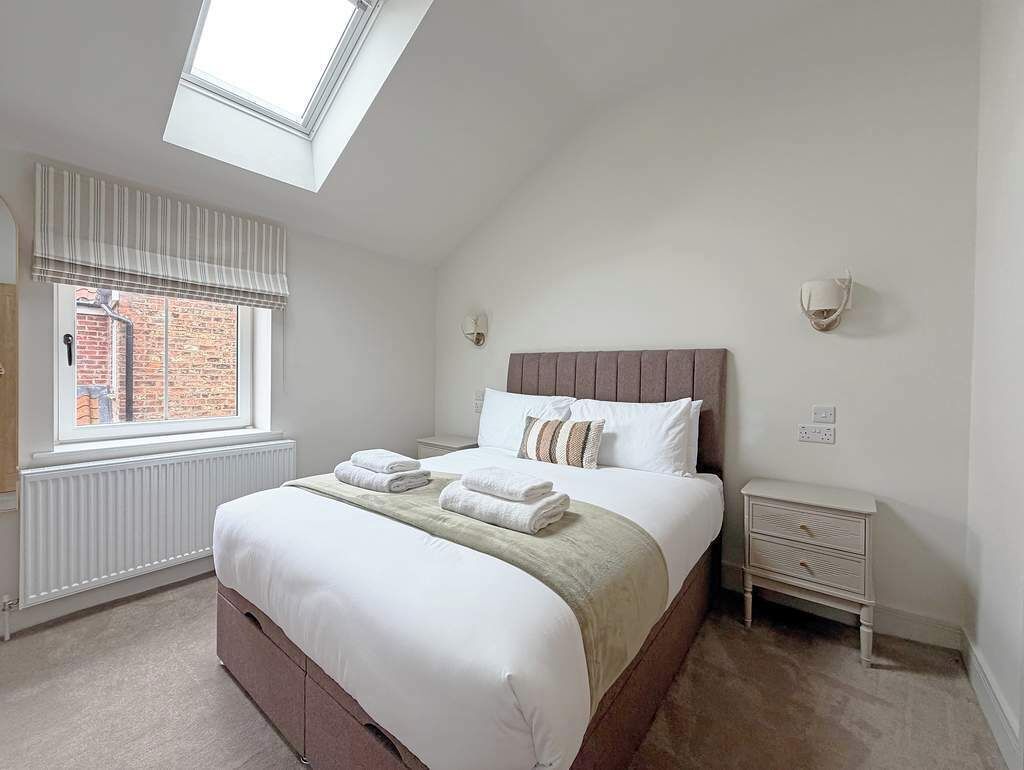 A bedroom with a skylight, a bed with pillows and a blanket, a bedside table, and a view of a brick building.