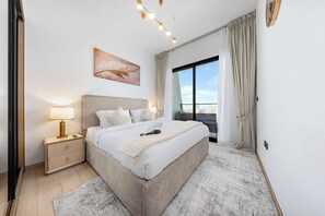 A modern bedroom with a large bed, a bedside table with a lamp, a window with curtains, and a city view. - Free Housekeeping Visits During Your Stay - Stayshort - 1 Bed Apartment in JVC That Sleeps 2 With Balcony (Dubai)