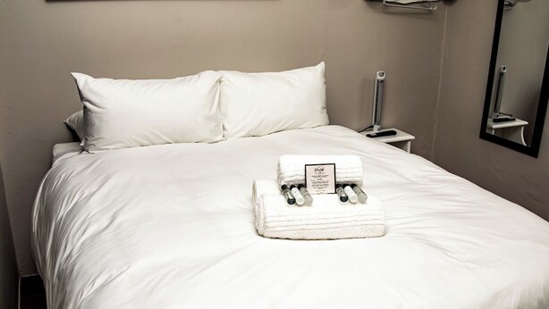 A neatly made bed with white linens, a folded towel, and a bottle of lotion on the bed.
