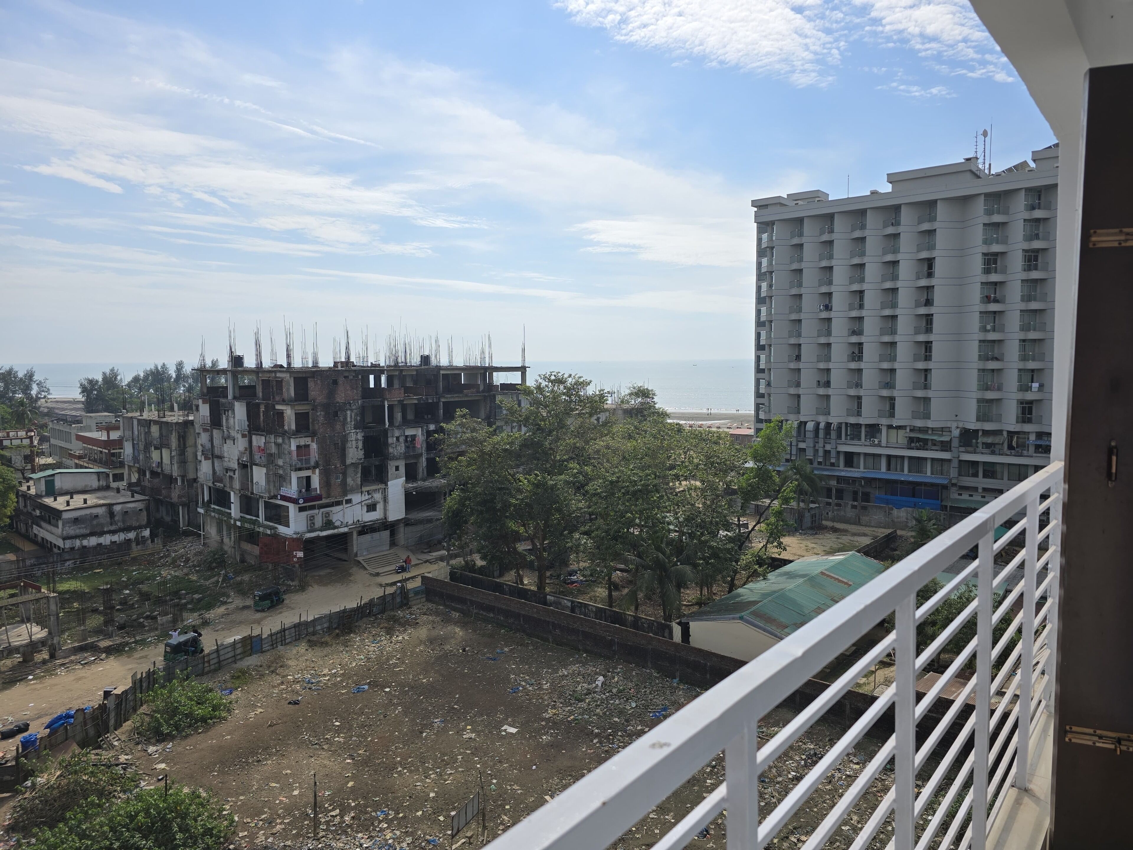 A view from a balcony overlooking a construction site with a building under construction, a beach, and a high-rise building in the background.