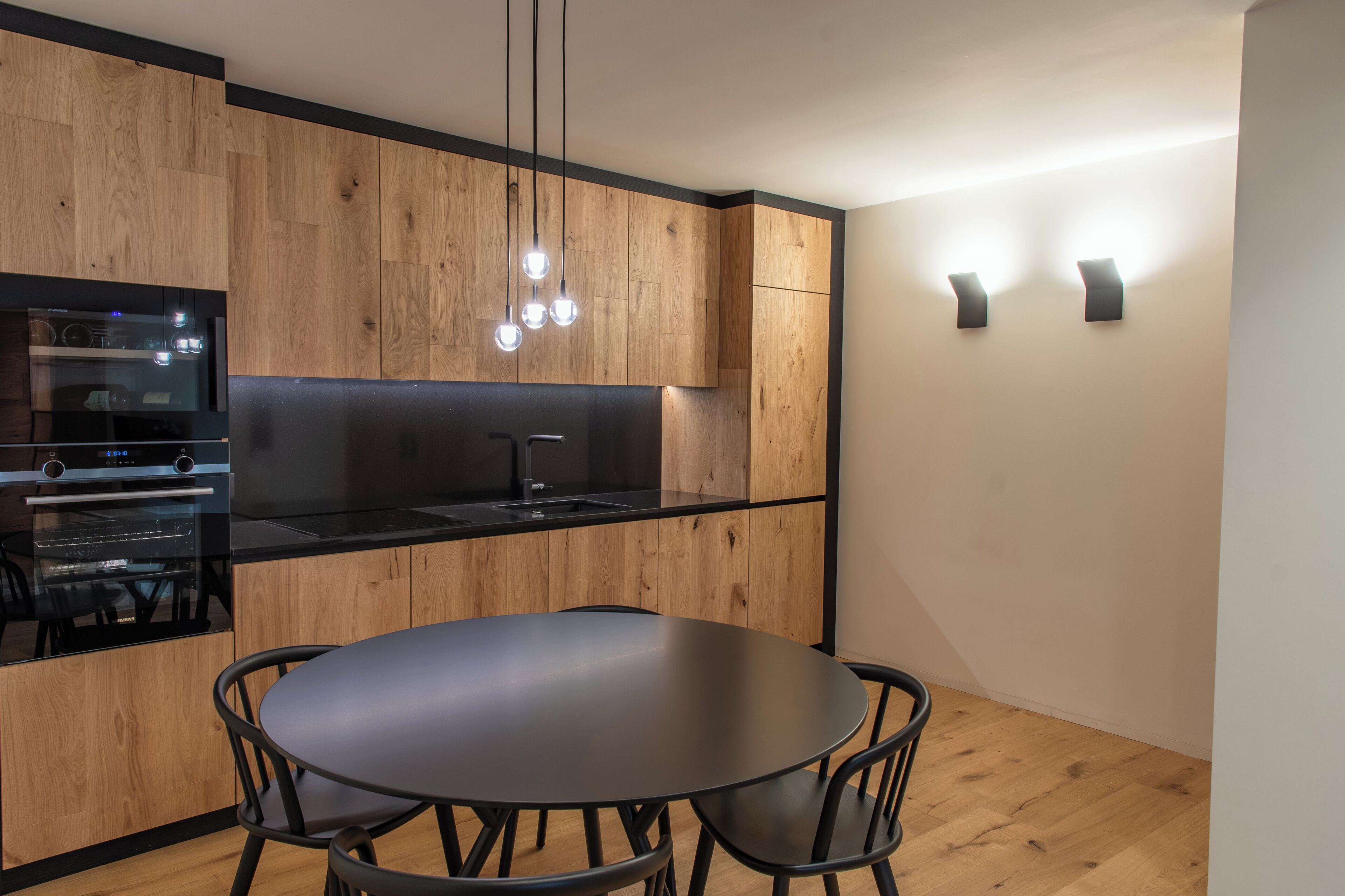 A modern kitchen with wooden cabinets, a black countertop, a round table with chairs, and pendant lights.