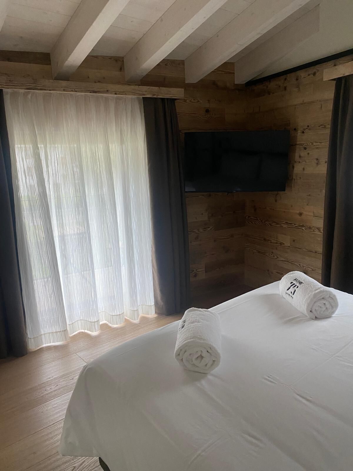 Quarto de hotel com teto de madeira, cama com roupa de cama branca, televisão na parede e janela com cortinas leves.