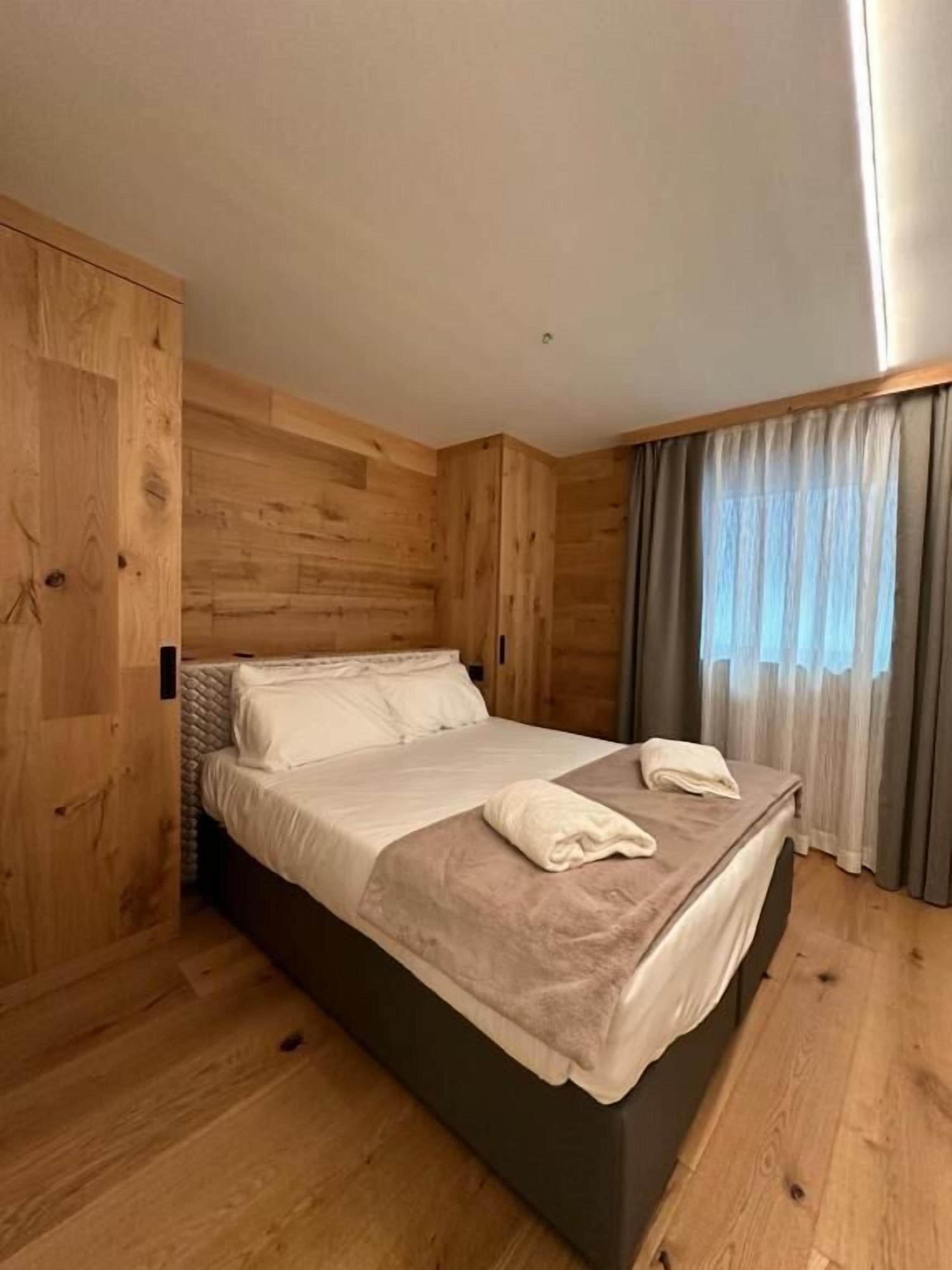 A room with a wooden floor, a bed with white linens, and a window with curtains.