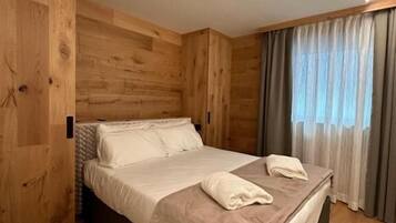 Una habitación con piso de madera, una cama con sábanas blancas y un ventanal con cortinas.