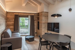 Una sala de estar moderna con un sofá, una mesa redonda y sillas, un televisor montado en la pared, y vistas al exterior a través de un ventanal grande.