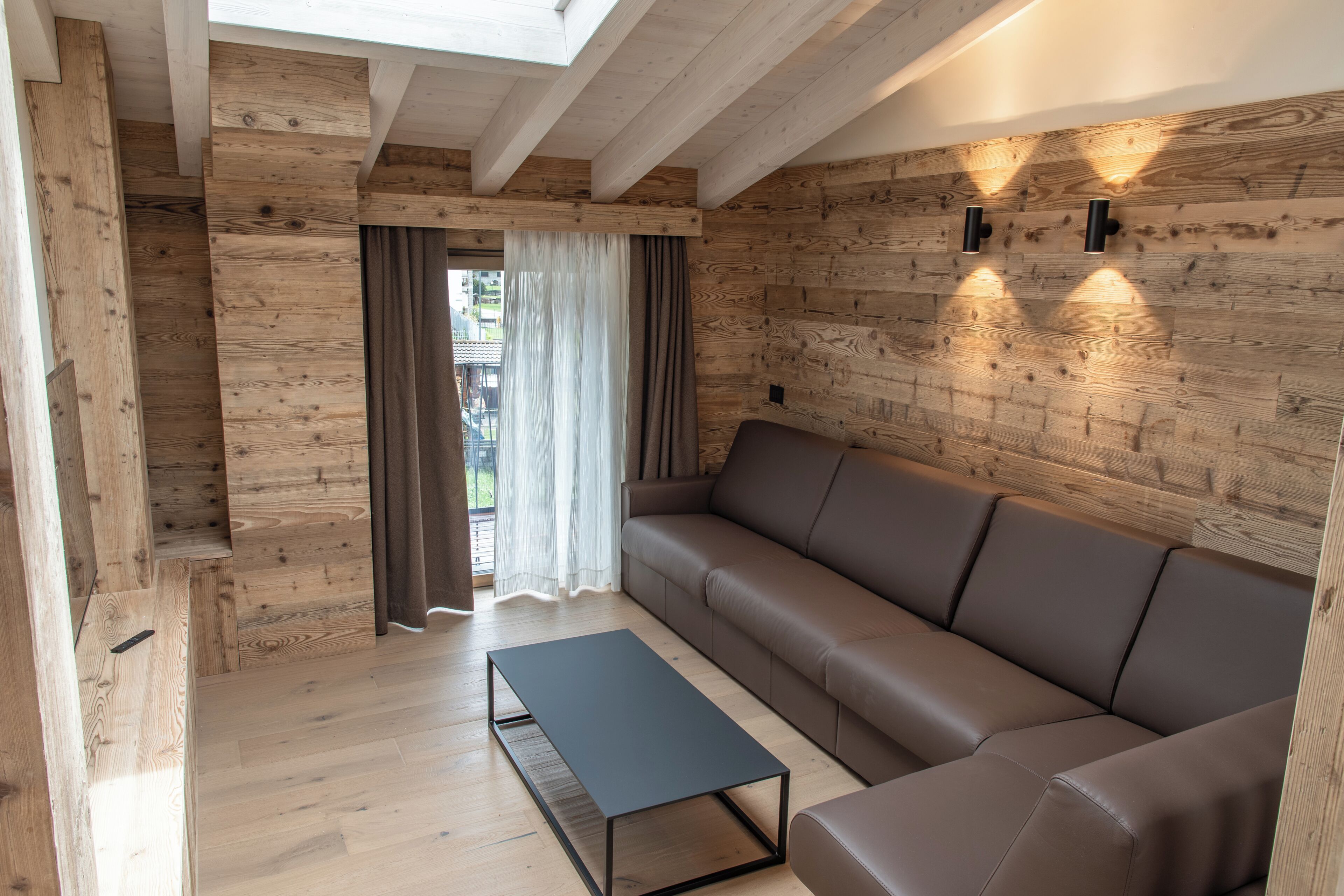 Una sala de estar moderna con techo de madera, un sofá de cuero marrón, una mesa de centro negra y vistas al exterior a través de una ventana con cortinas translúcidas.