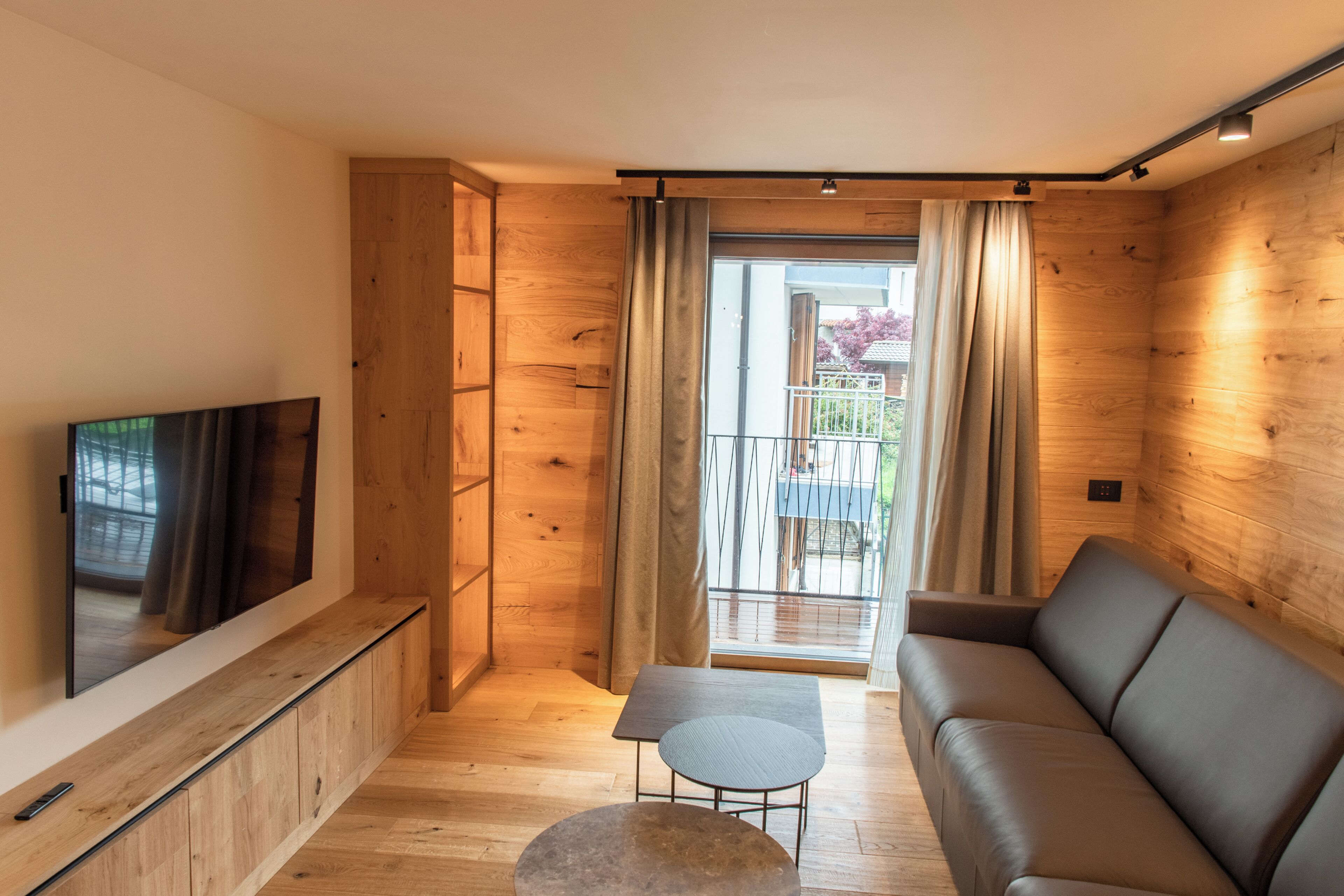 A modern living room with a flat-screen TV, wooden walls, a sofa, a small table, and a sliding glass door leading to an outdoor balcony.