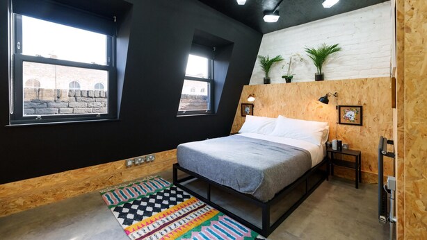 A bedroom with a bed, two windows, a wooden headboard, a nightstand, a lamp, and a colorful rug.