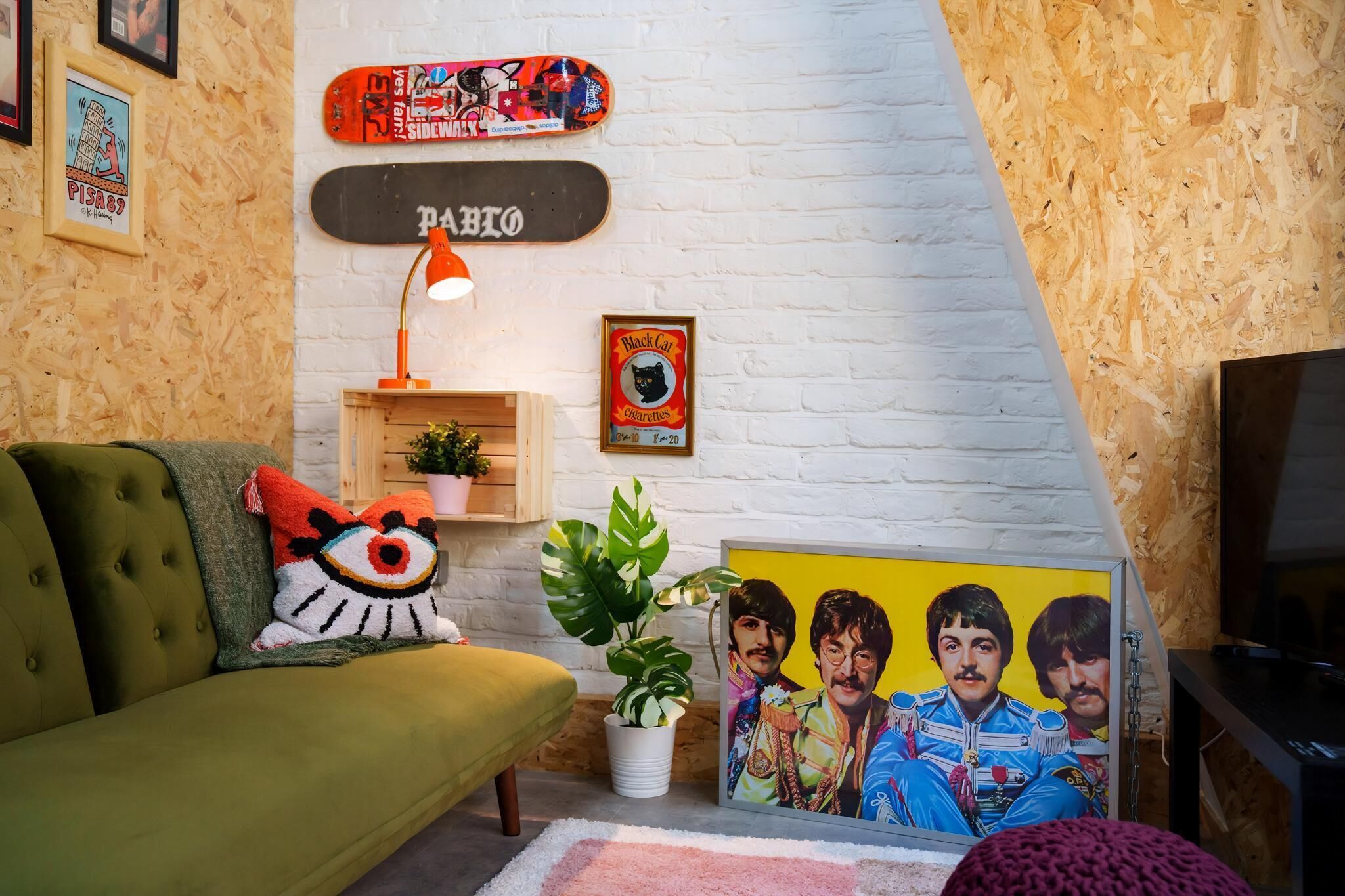 A living room with a green sofa, a white brick wall, a skateboard, a framed poster, a plant, and a TV.