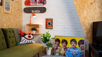 A living room with a green sofa, a white brick wall, a skateboard, a framed poster, a plant, and a TV.