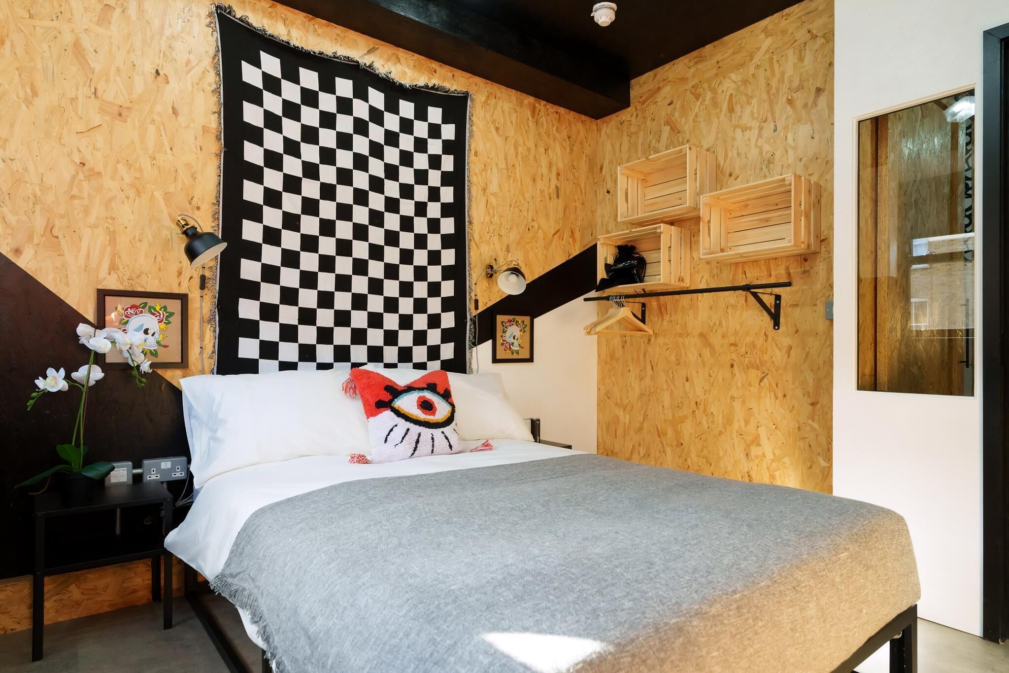 Ein Schlafzimmer mit karierter Wand, einem Bett mit grauer Decke, einem an der Wand montierten Regal mit Holzschachteln und einem gerahmten Bild auf dem Nachttisch.