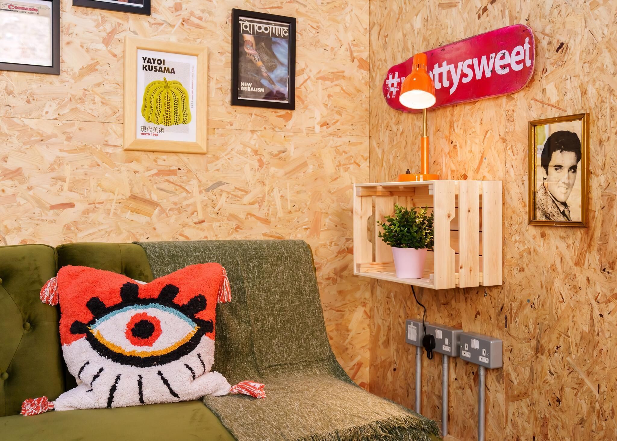 A room with a green sofa, a colorful eye-shaped pillow, a wooden shelf with a plant, and a wall-mounted sign.