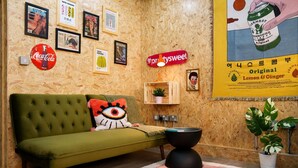 A green sofa with a cushion, a wall with framed pictures, a Coca-Cola sign, a shelf with a plant, and a poster advertising lemon & ginger beer.